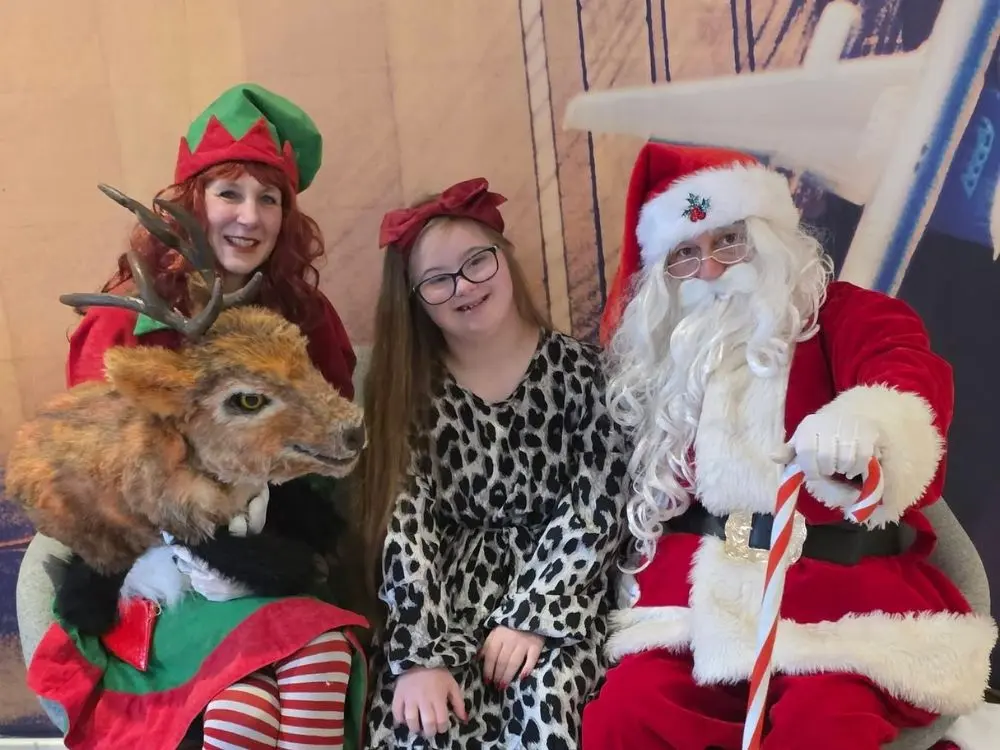 A young girl with a bow in her hair posing for a photo with Santa and an Elf at Hampton by Hilton.