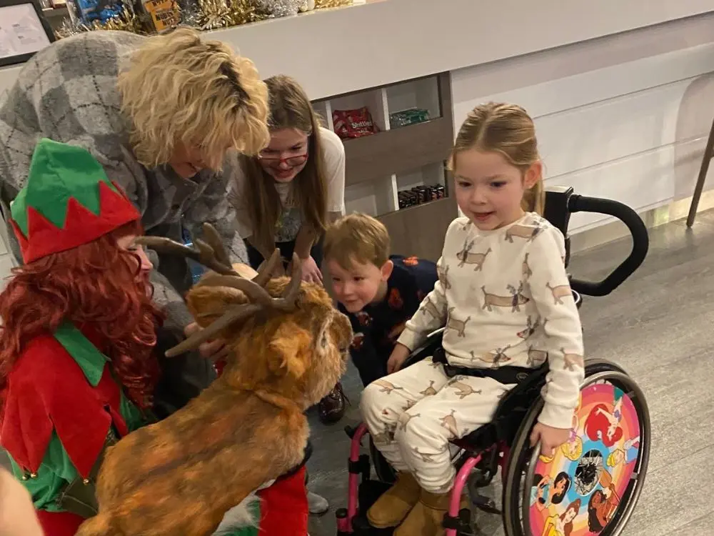 A child in a wheelchair interacts with a realistic reindeer puppet held by a festive Elf.