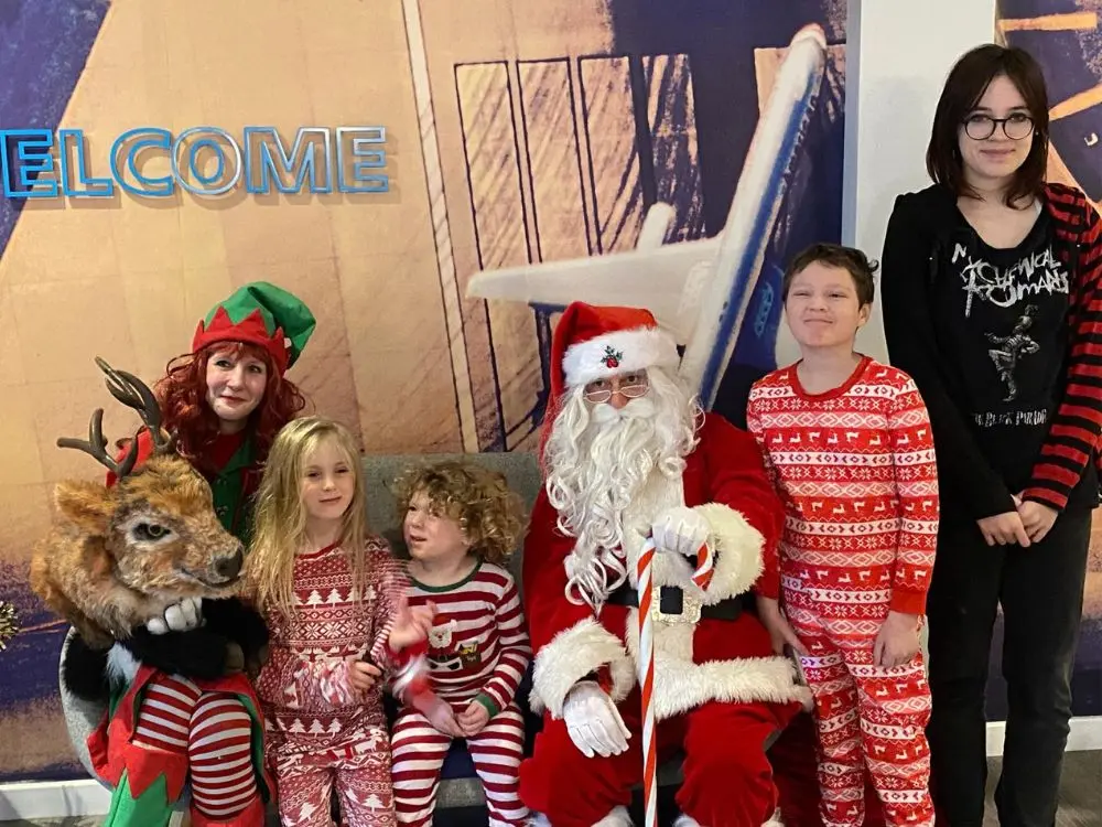 A group of children in festive pajamas posing with Father Christmas, an Elf, and a reindeer puppet.