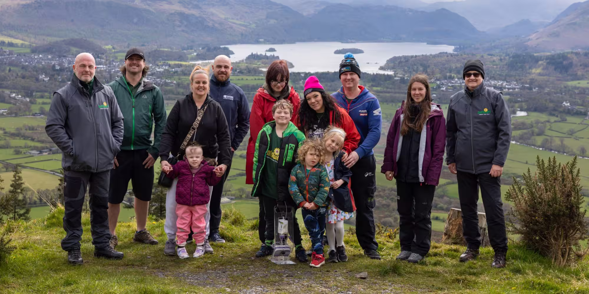 Sunflowers families and volunteers standing together on a hillside at Calvert Lakes, with Derwentwater and the surrounding fells visible in the background
