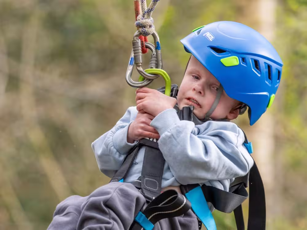 A young child in a blue helmet and harness on a zip wire at Calvert Lakes, concentrating as they hold on