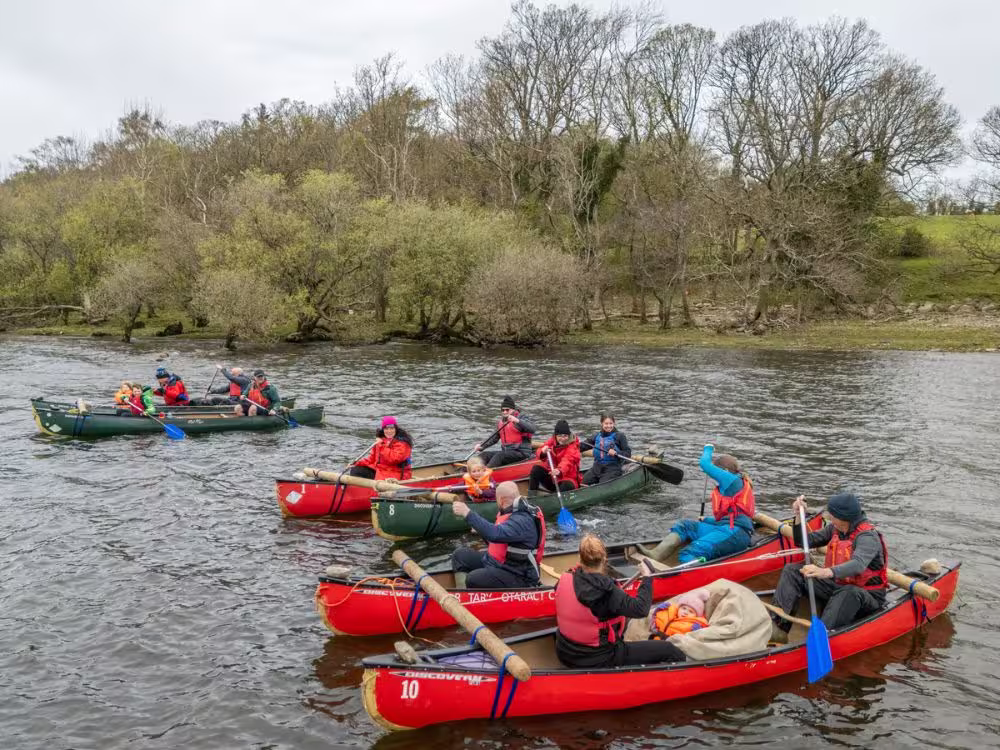 A group of Sunflowers families and volunteers canoeing together on the river at Calvert Lakes, wearing life jackets and paddling red canoes