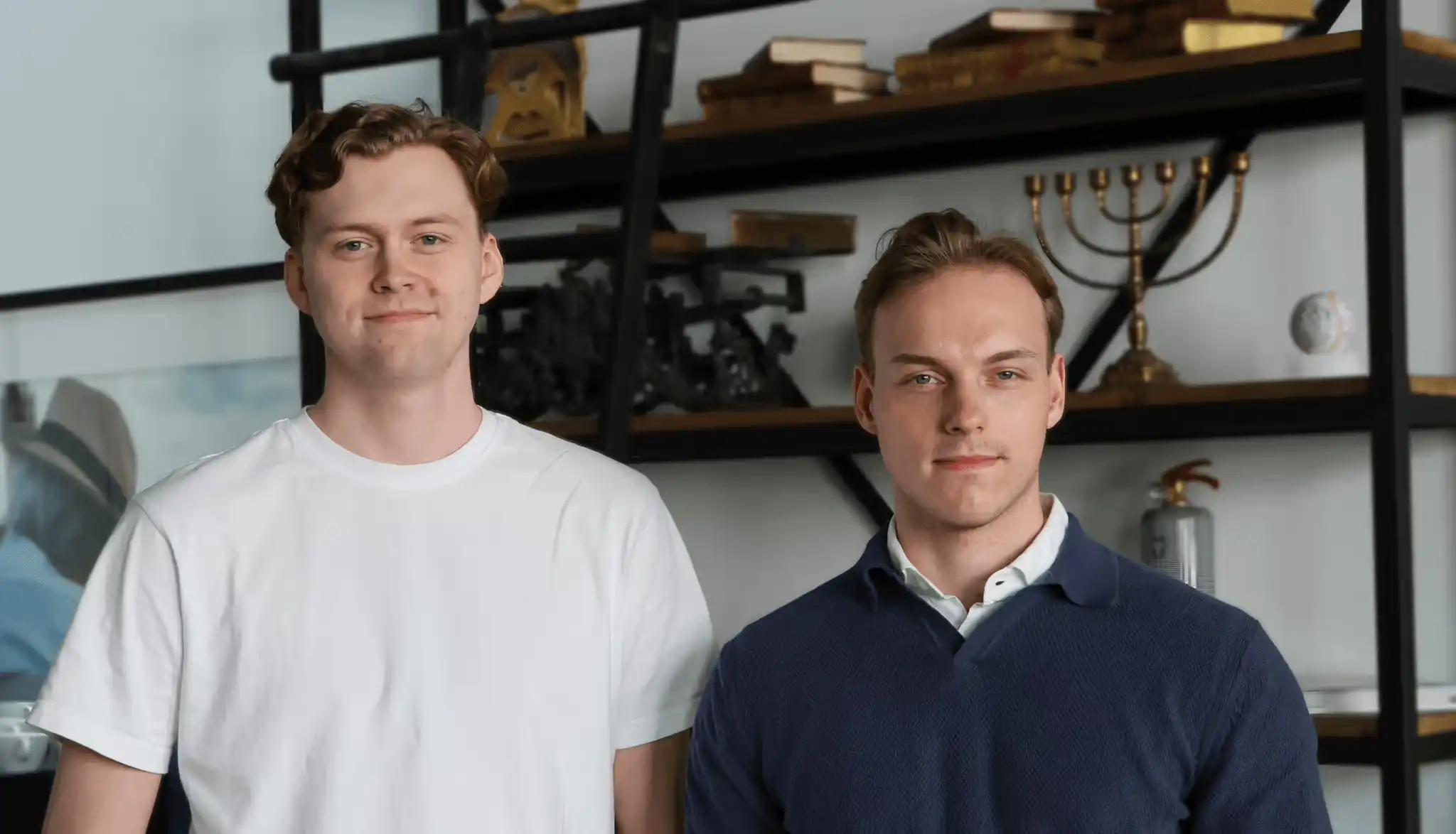 Två män står framför en bokhylla och små ler med stängd mun.