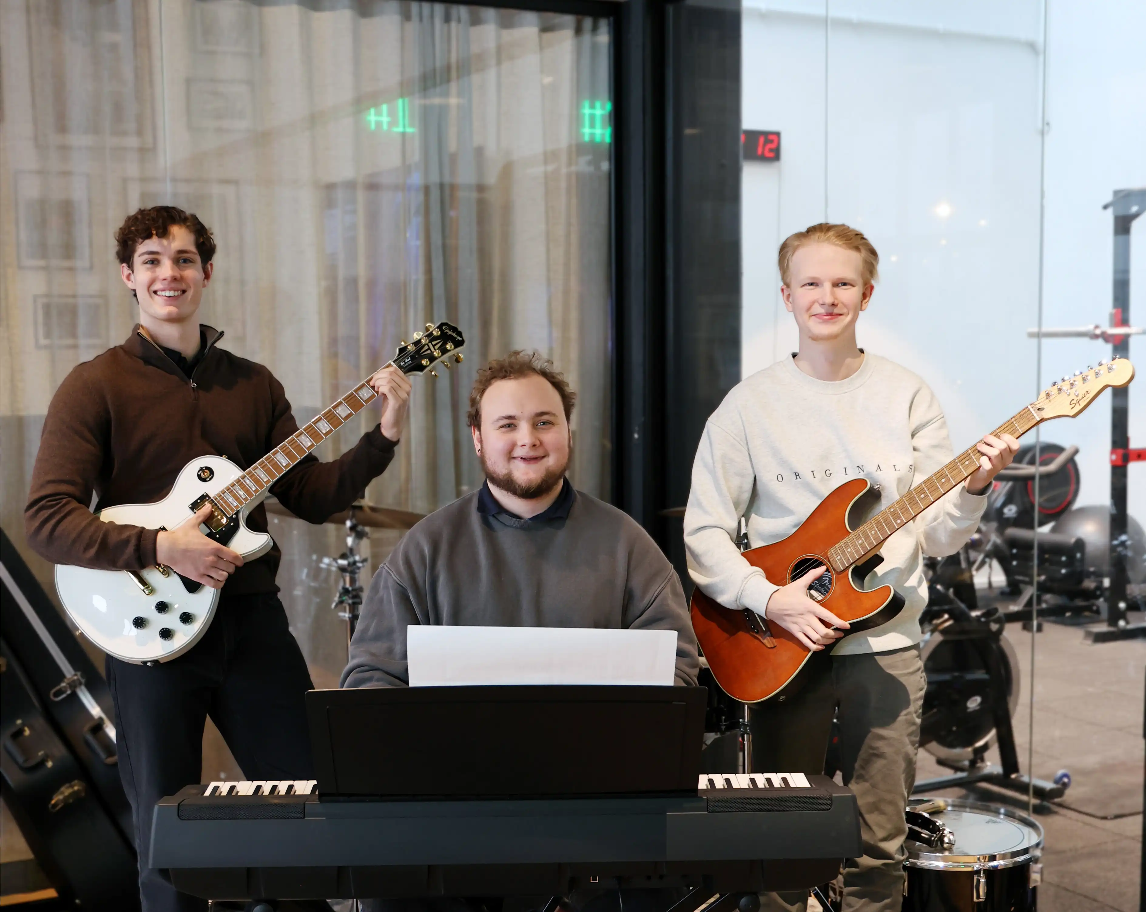 Tre unga män står i ett musikrum. Två av dem håller elgitarrer och ler mot kameran – den ena med en vit gitarr till vänster och den andra med en brun gitarr till höger. Den tredje personen sitter i mitten vid ett elpiano med noter framför sig. I bakgrunden syns trumset, gitarrfodral och gymutrustning bakom glasväggar.