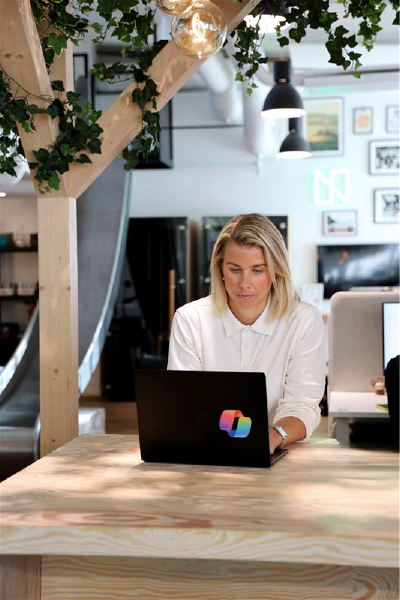 En kvinna sitter vid ett ljust träbord i ett modernt kontorslandskap och arbetar fokuserat på en laptop med färgglad logotyp. Gröna växter och varm belysning ramar in miljön, som har en skandinavisk och kreativ känsla.