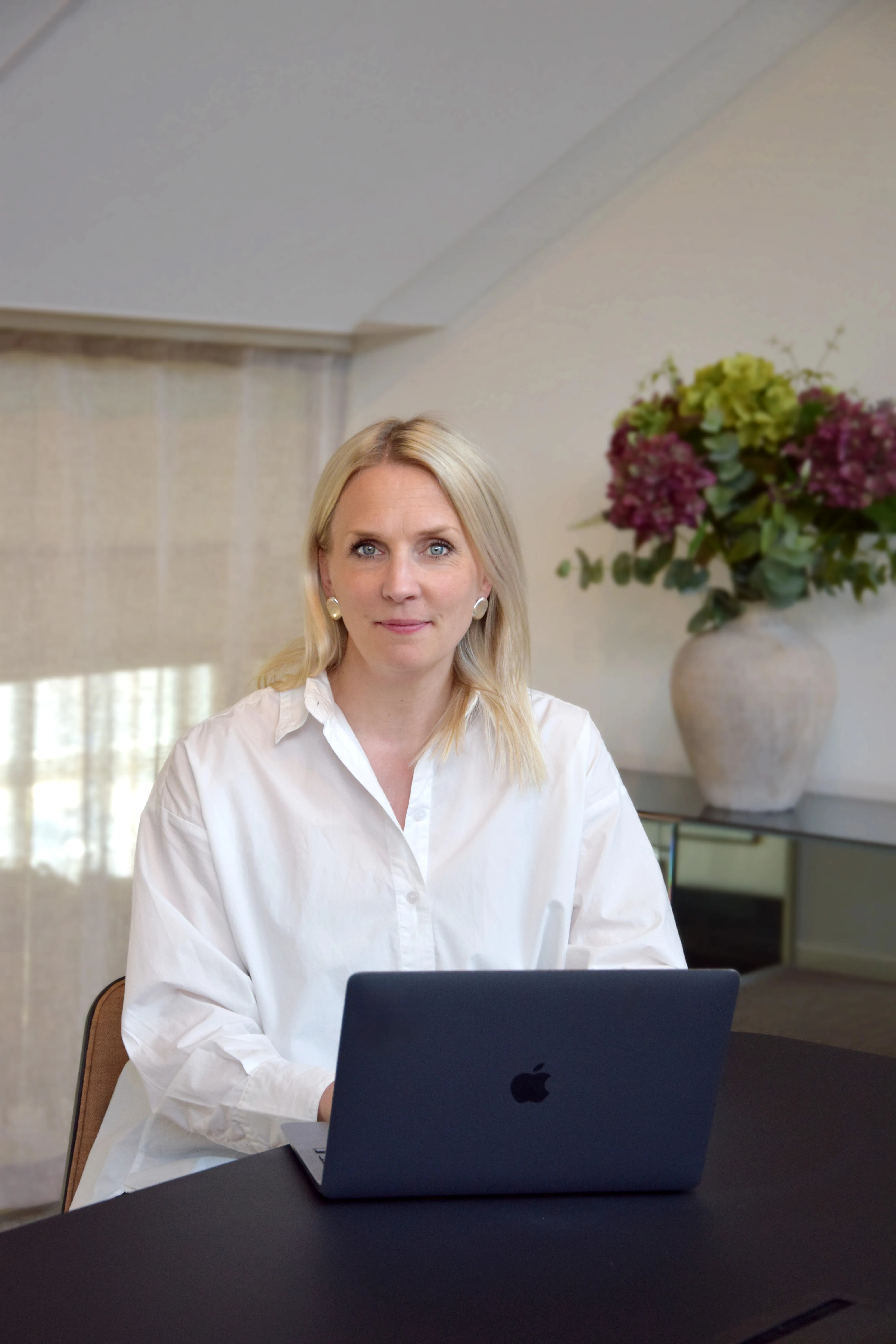 Kvinna med ljust hår sitter vid ett bord med en mörk laptop och tittar in i kameran. Hon bär en vit skjorta och sitter i ett ljust rum med en stor vas med blommor i bakgrunden.