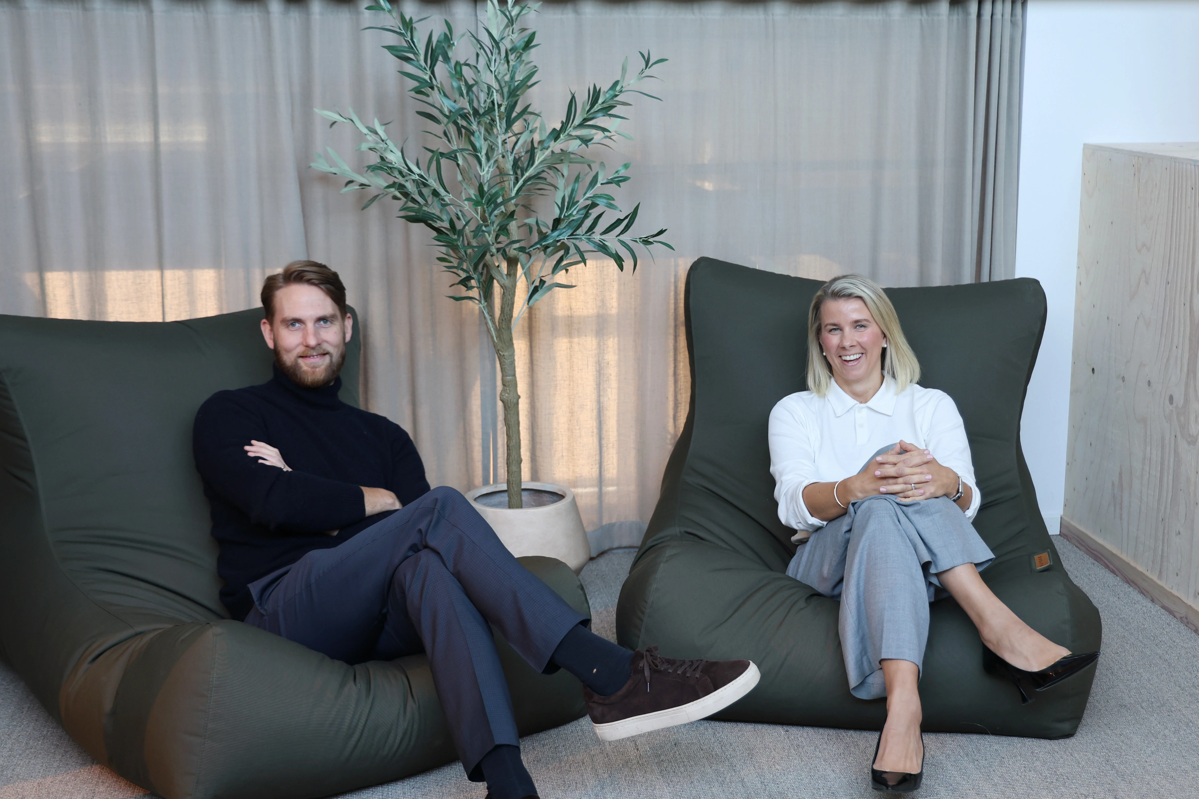 En kvinna och en man sitter bredvid varandra på en grön träbänk i ett modernt kontor. Bakom dem syns en glasvägg med Norteams logotyp i vitt, samt inslag av kontorsinredning och ljus från fönster. Kvinnan bär en vit skjorta och grå byxor, mannen en mörk tröja och grå byxor. Båda har händerna vilande i knät.