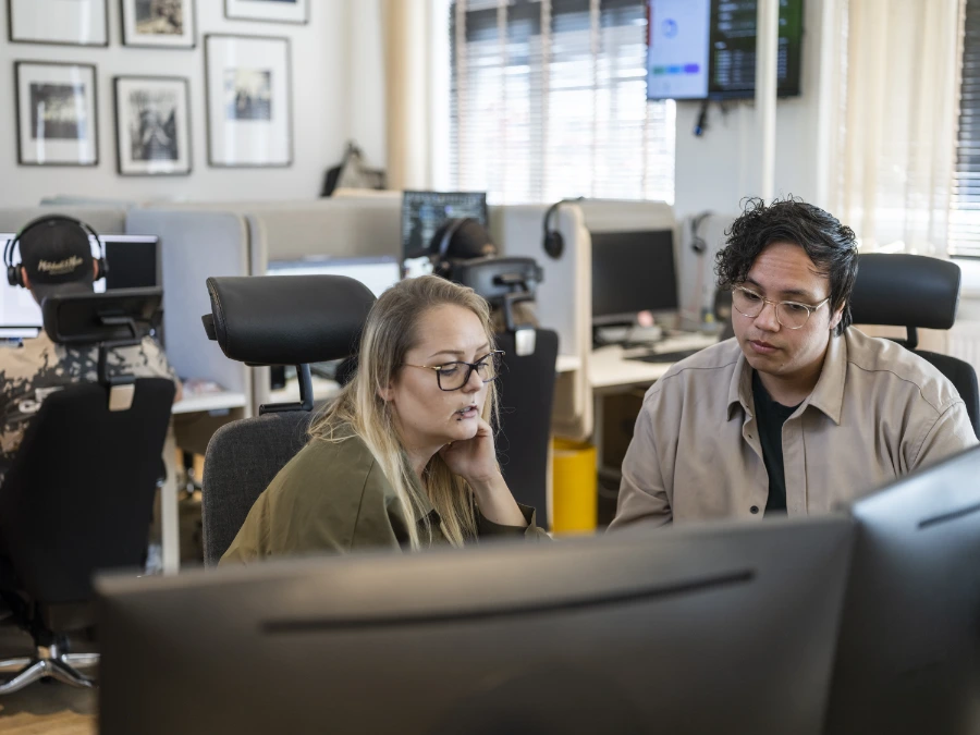Två kollegor sitter vid ett kontor och samarbetar framför en datorskärm, medan andra medarbetare arbetar i bakgrunden i ett öppet kontorslandskap.