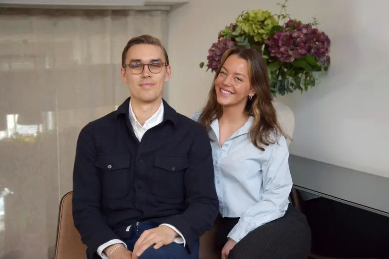 En man och en kvinna sitter bredvid varandra inomhus och ler mot kameran. Mannen har har glasögon, kort kammat hår och bär en mörk jacka över en vit skjorta. Kvinnan har långt brunt hår och bär en ljusblå skjorta och ler brett. I bakgrunden syns en stor bukett med lila och gröna hortensior i en vit vas.