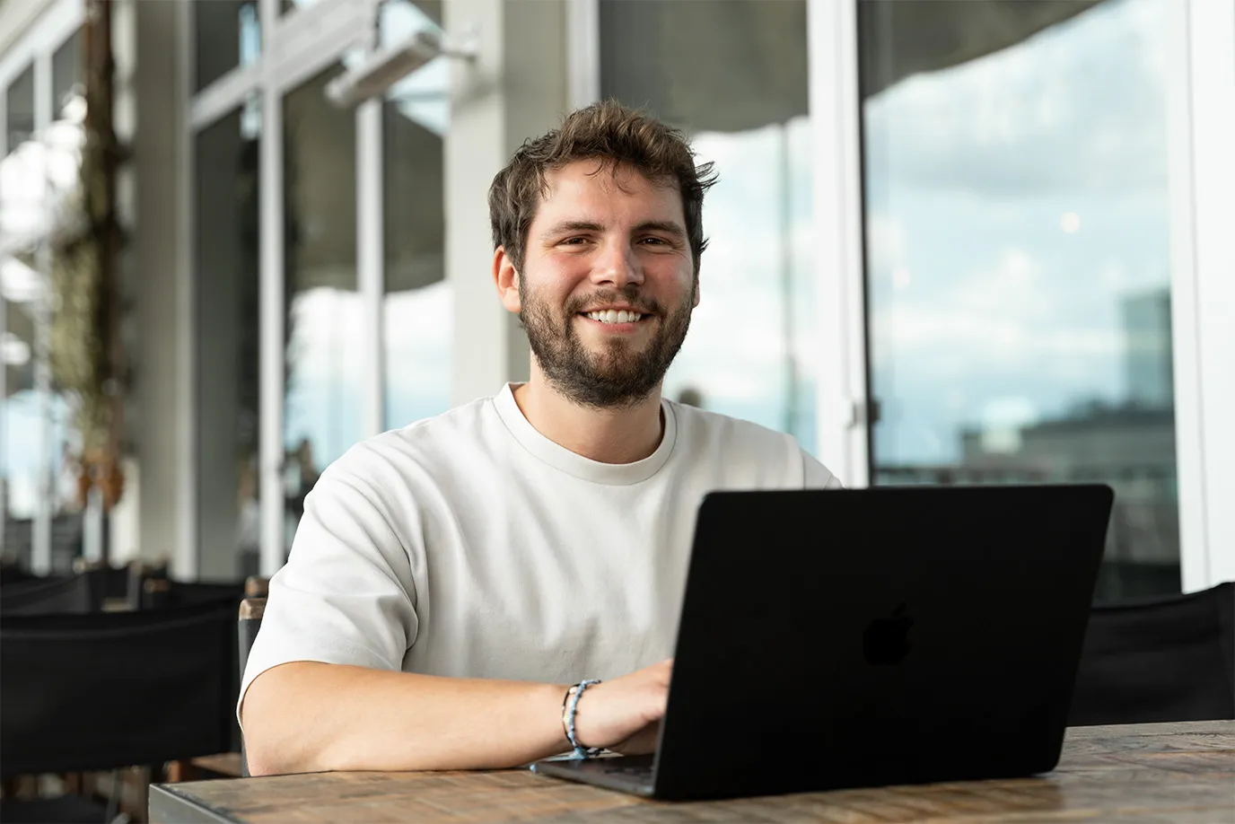 Nils sitzt draußen vor einem Laptop und lächelt in die Kamera