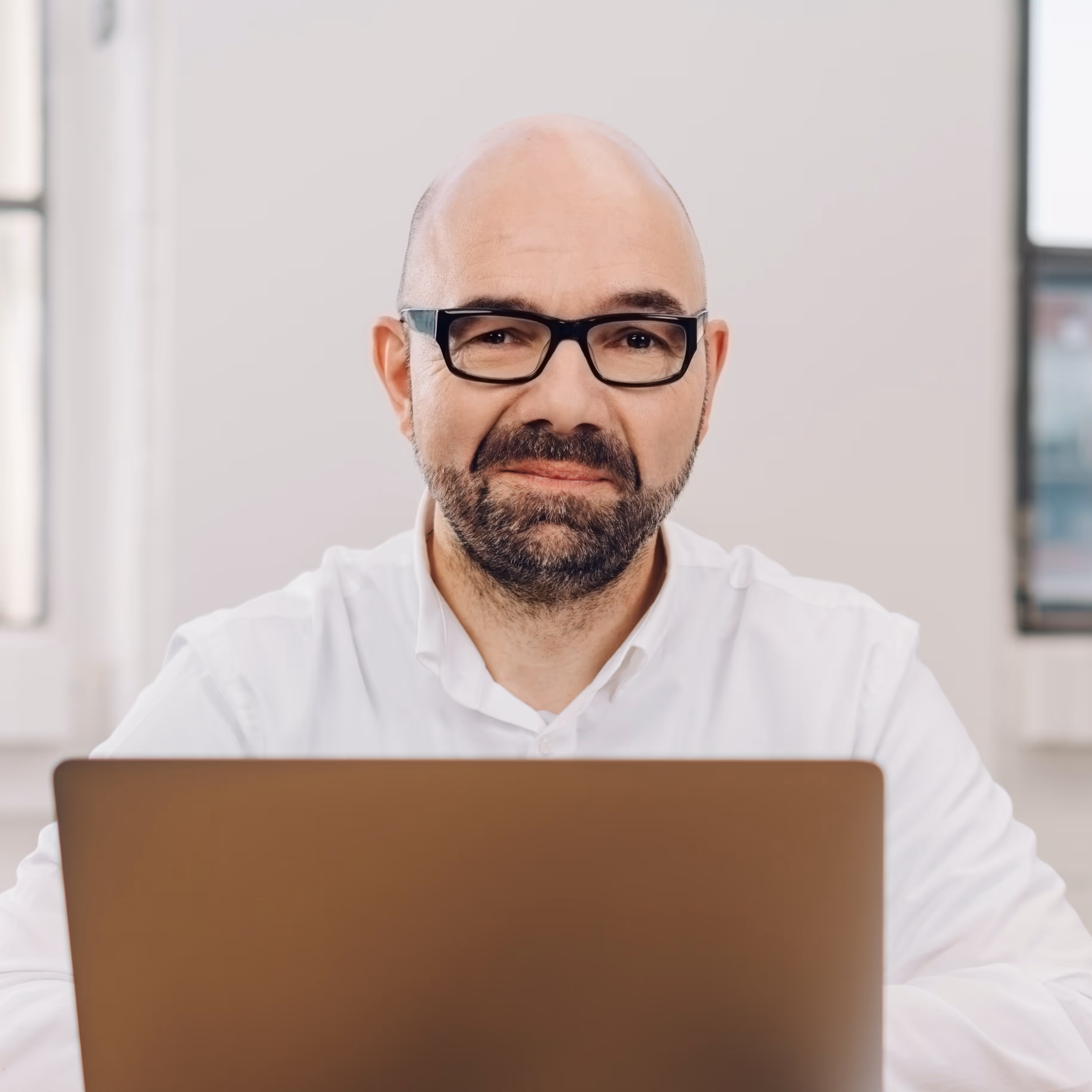 Weißer Mann mittleren Alters mit Glatze, Vollbart und Brille sitzt vor einem Laptop und lächelt leicht in die Kamera