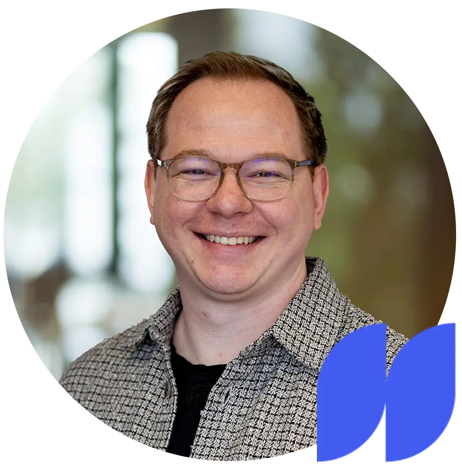 Lächelnder Mann mit roten Haaren, Brille und weißem Hemd vor grauem Hintergrund.