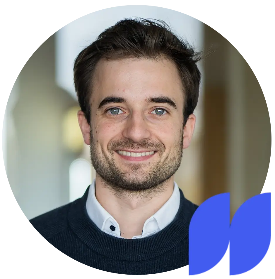 Junger Mann mit braunem Haar, Bart und blauem Pullover lächelt vor grauem Hintergrund.