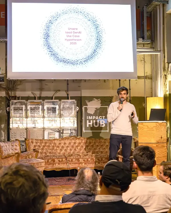 Ein Mann hält eine Präsentation vor Publikum in einem Raum mit Vintage-Möbeln und einem Bildschirm, der den Text 'Unsere toad GenAI Use Case Hypothesen 2025' zeigt.