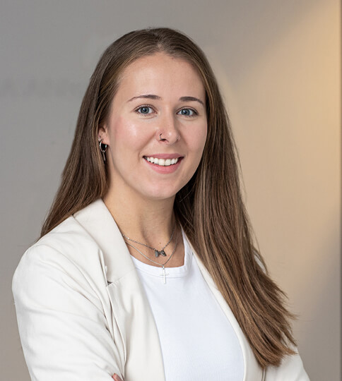 Junge Frau mit langen braunen Haaren, die einen weißen Blazer und weiße Bluse trägt, lächelt vor grauem und beigem Hintergrund.