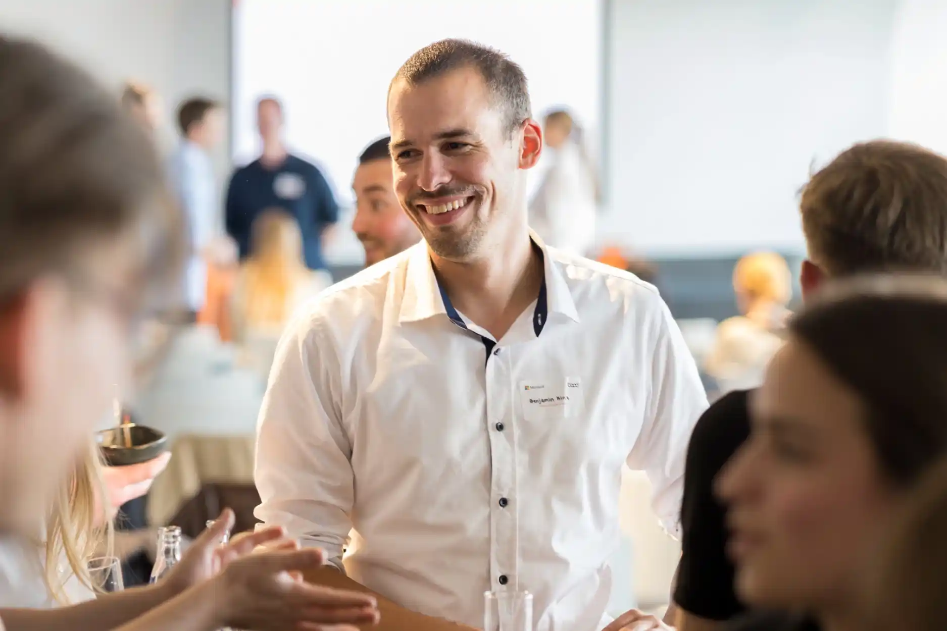Lächelnder Mann in weißem Hemd unterhält sich auf einer Veranstaltung mit anderen Menschen.
