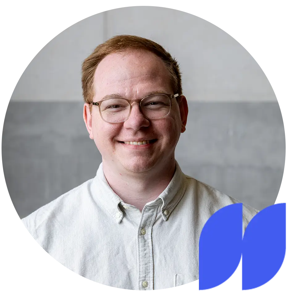 Lächelnder Mann mit roten Haaren, Brille und weißem Hemd vor grauem Hintergrund.