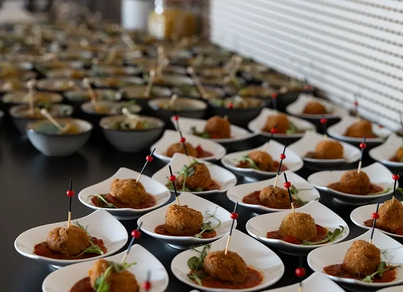 Zwei Männer sitzen an einem Tisch vor einem Laptop, einer hält eine grüne Flasche, im Hintergrund Fenster mit Blick auf Gebäude.