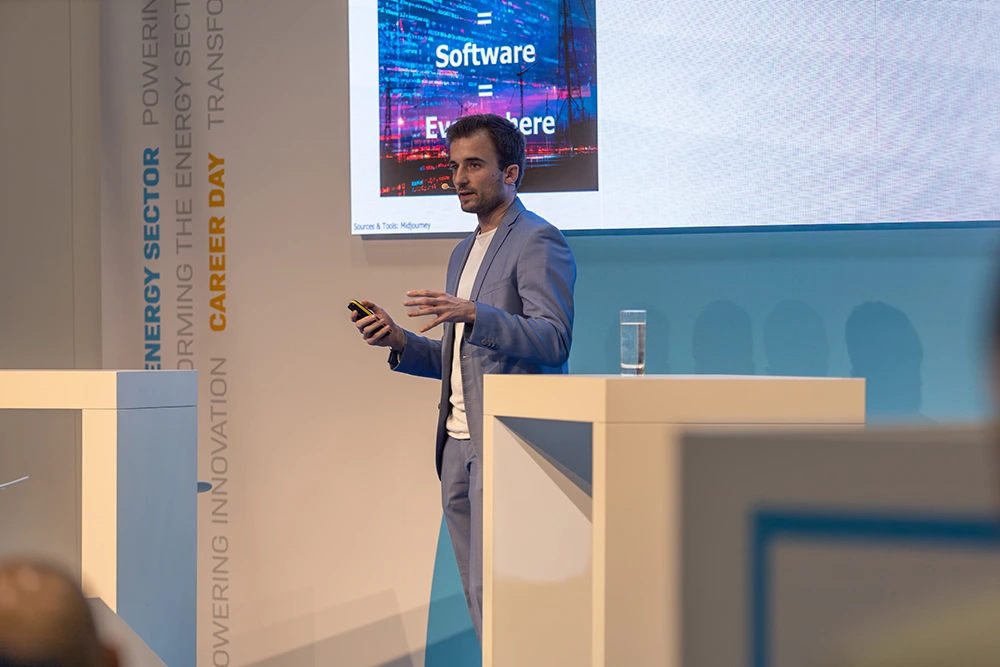 Zwei Männer sitzen an einem Tisch vor einem Laptop, einer hält eine grüne Flasche, im Hintergrund Fenster mit Blick auf Gebäude.