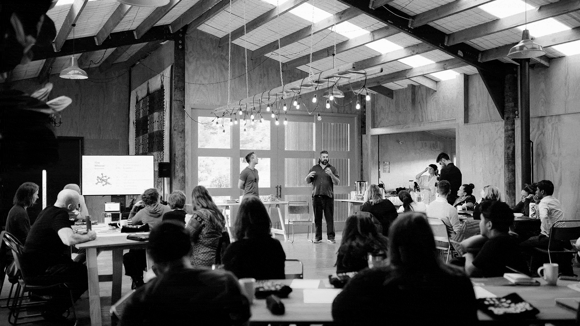Two men standing and speaking in front of an audience seated at tables in a spacious, rustic room with exposed beams and hanging lights.