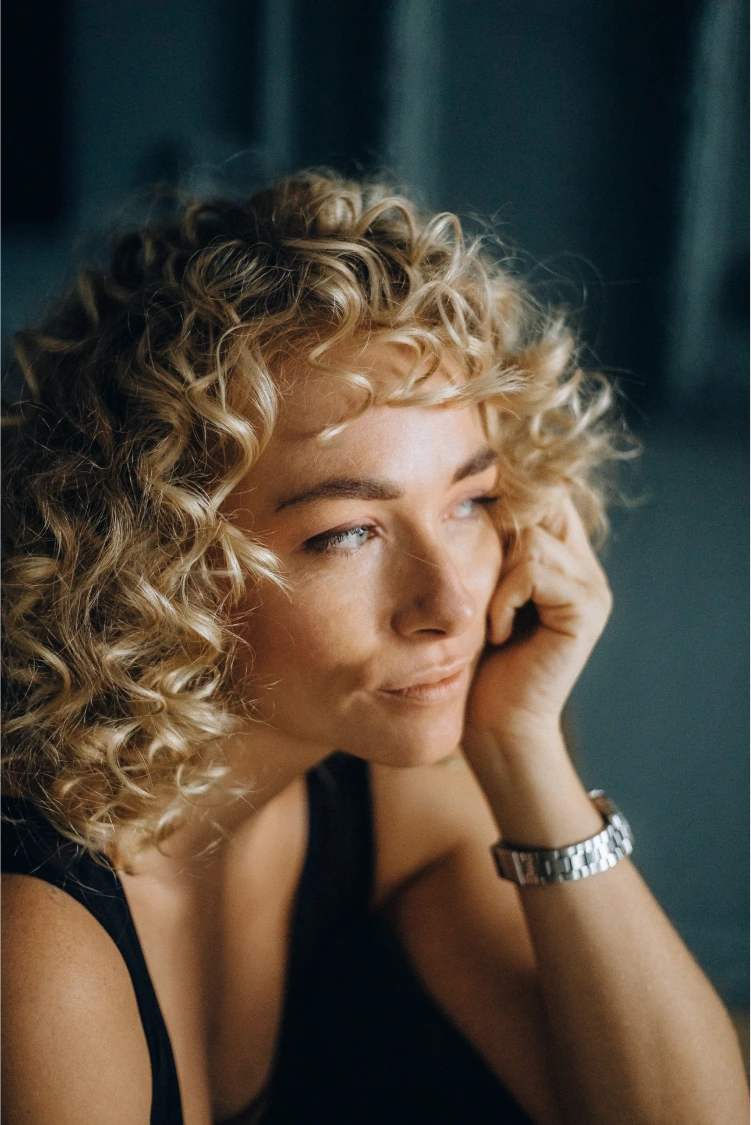 A woman with curly blonde hair, resting her head on her hand, gazing thoughtfully into the distance.