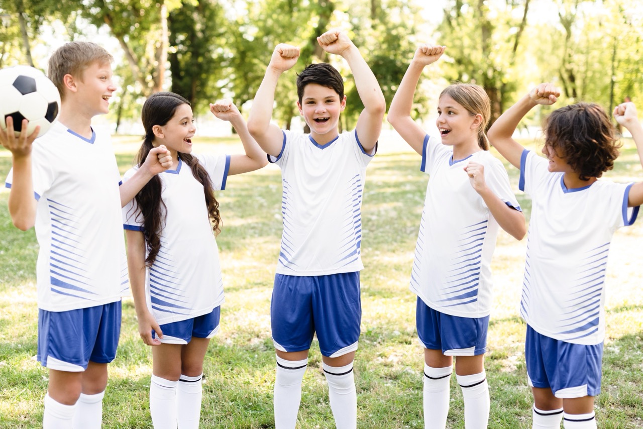 Five children in white and blue soccer uniforms cheering outdoors on grass with trees in the background.