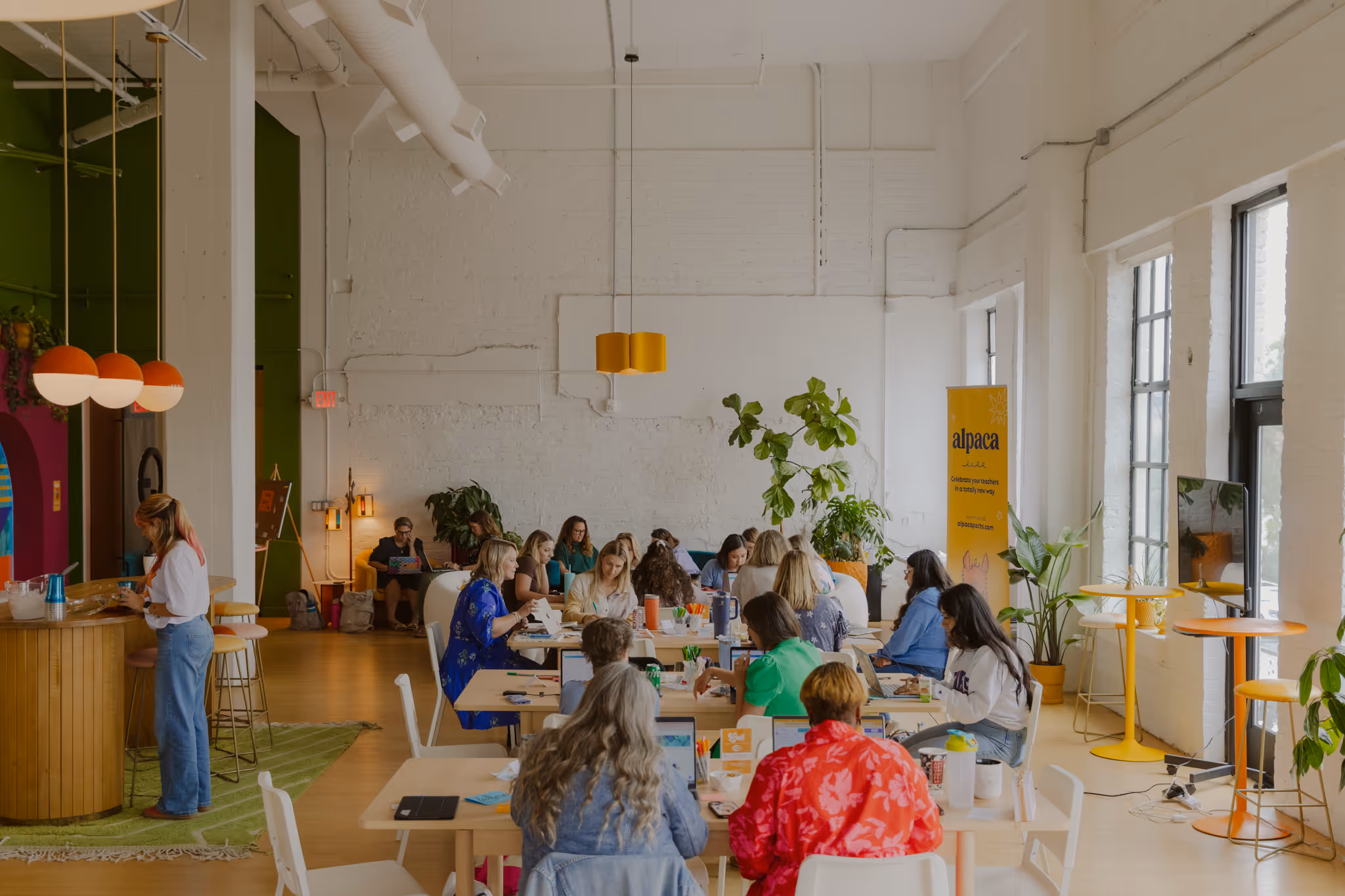 Group of people seated at tables working on laptops in a bright, modern coworking space with plants and a yellow Alpaca banner.