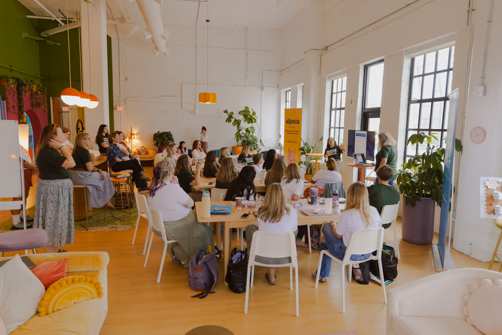 Group of people seated and standing in a bright room attending a presentation with a yellow alpaca banner and large windows.