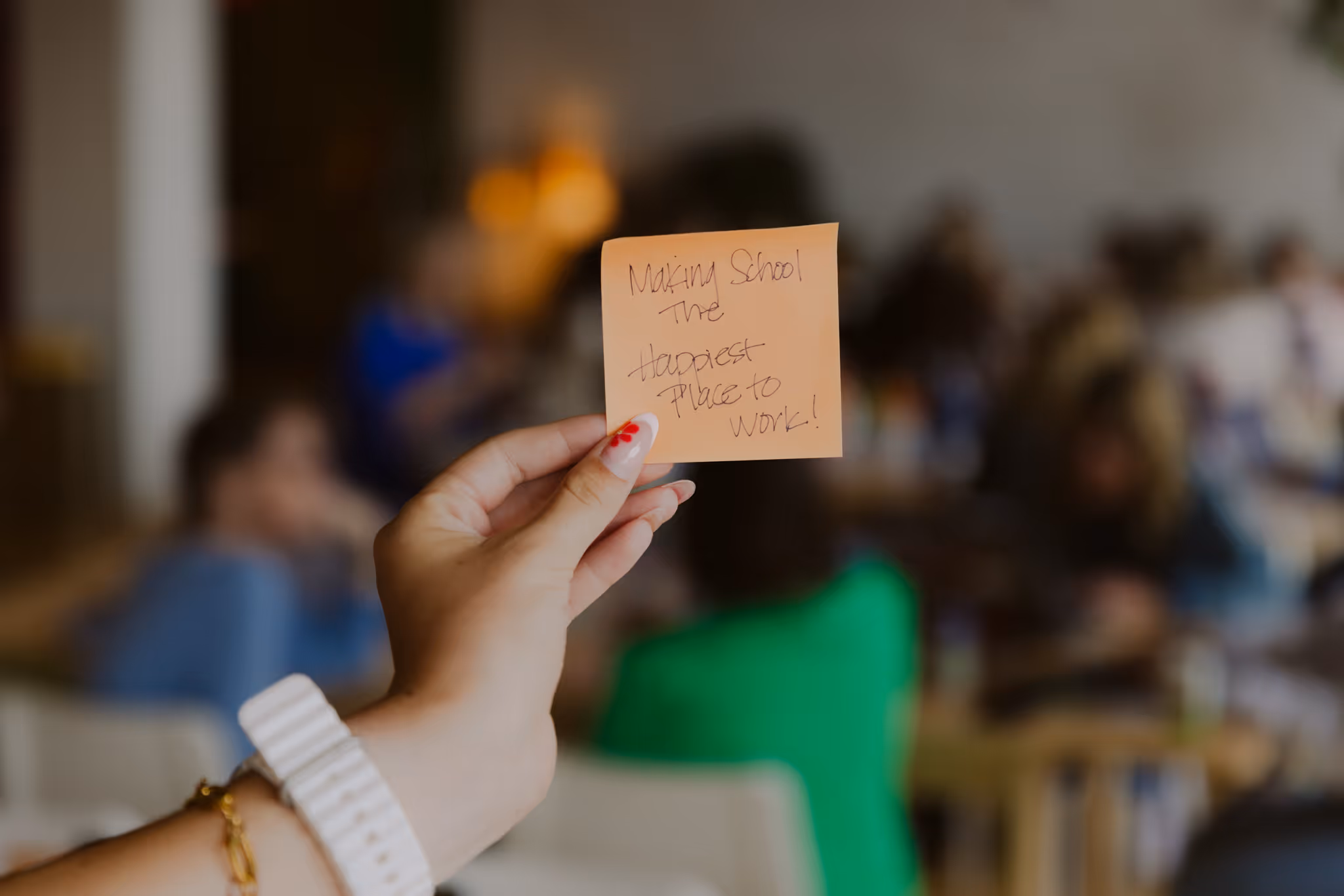 Hand holding an orange sticky note with the handwritten message: Making School The Happiest Place to Work!