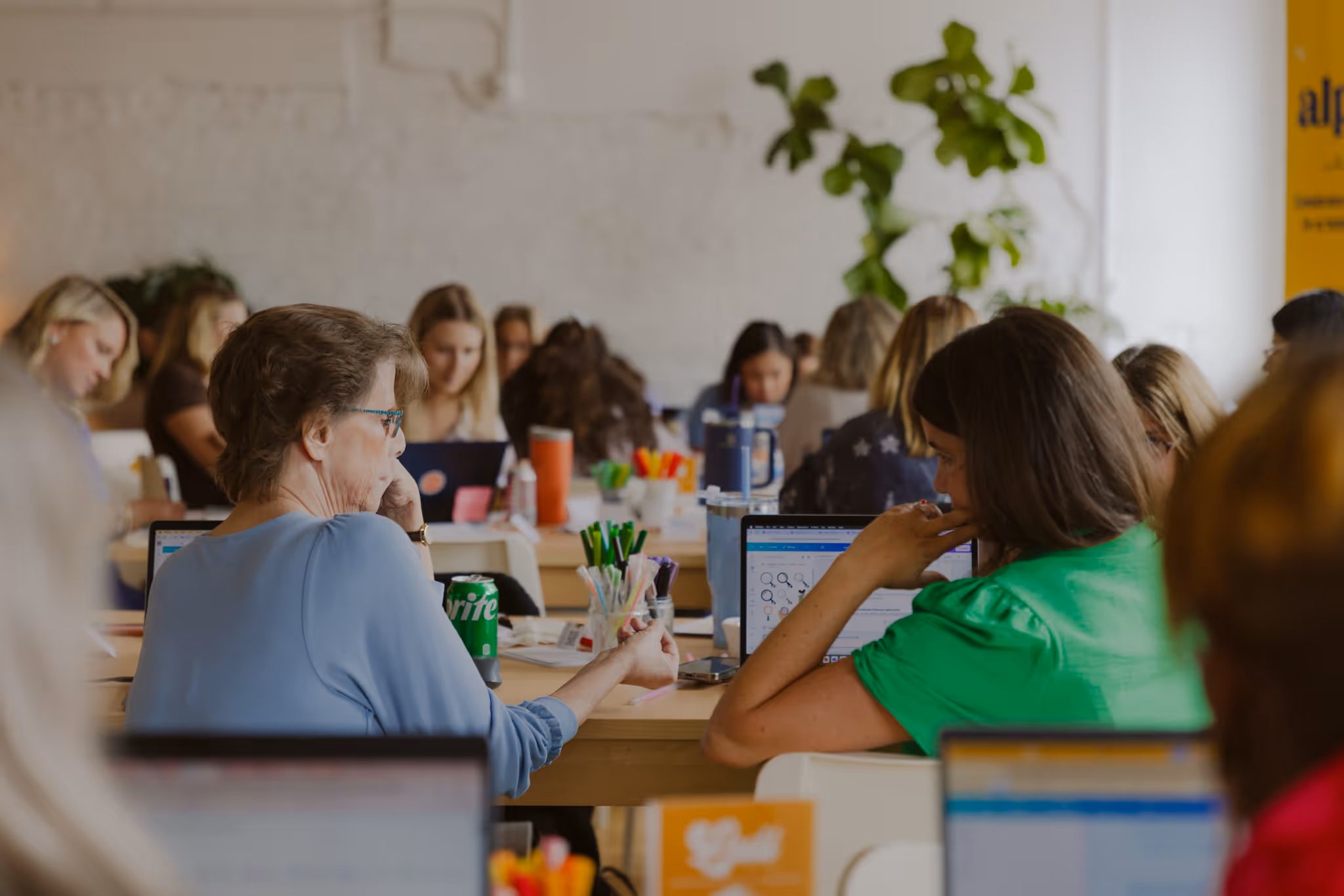 Group of people working on laptops at a large shared table in a bright, open office space.