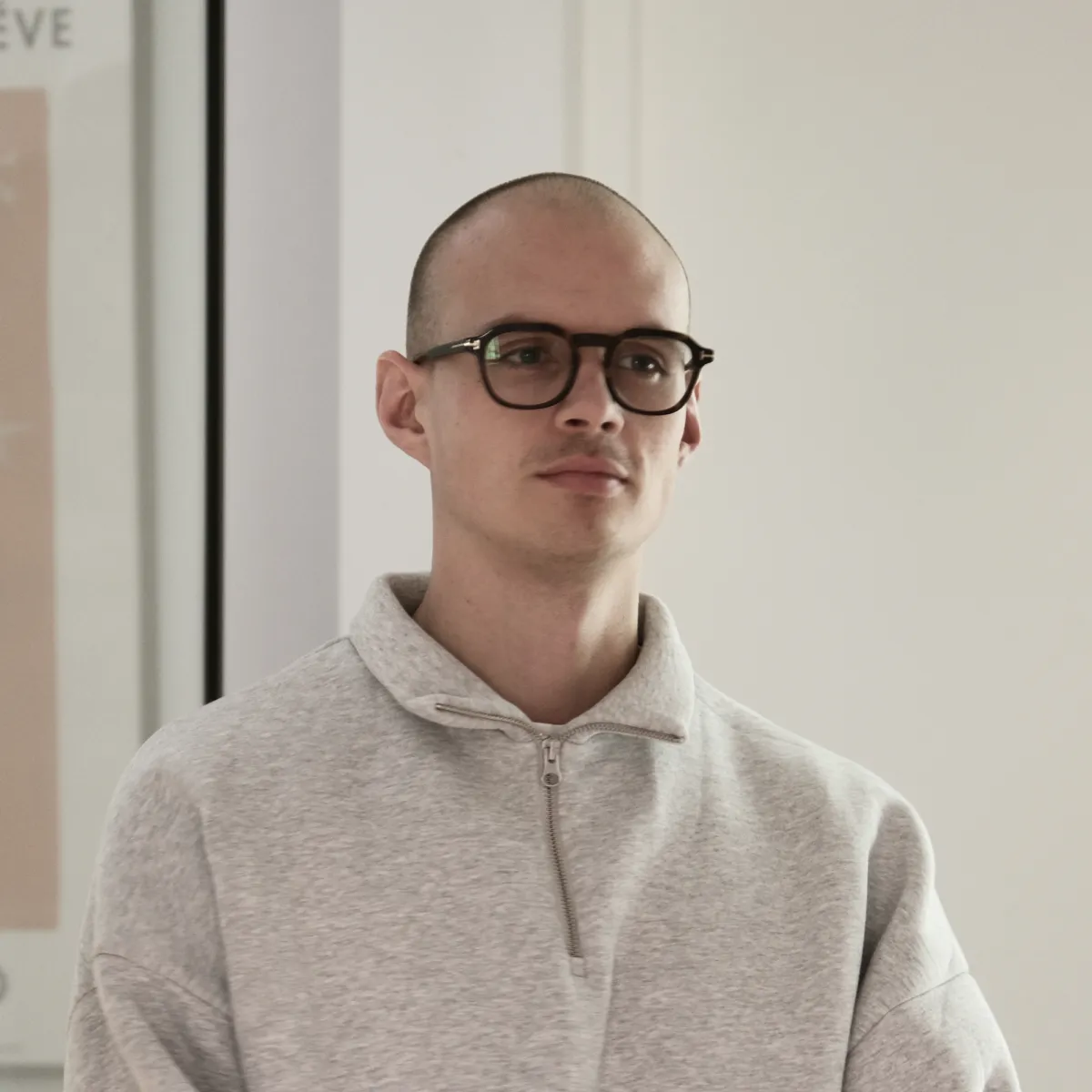 Jonge man, Leander Haesaert, met een grijze trui en een bril, staand in een neutrale binnenruimte.