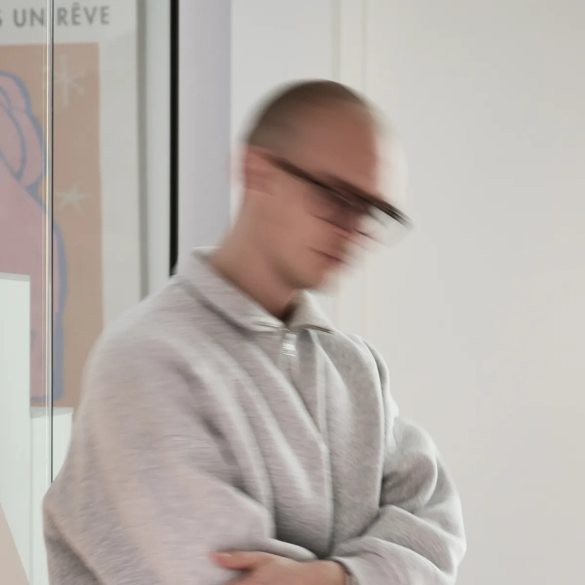 Vage afbeelding van een persoon met een grijze trui en zwarte bril, met armen gekruist, staand tegen een witte muur.