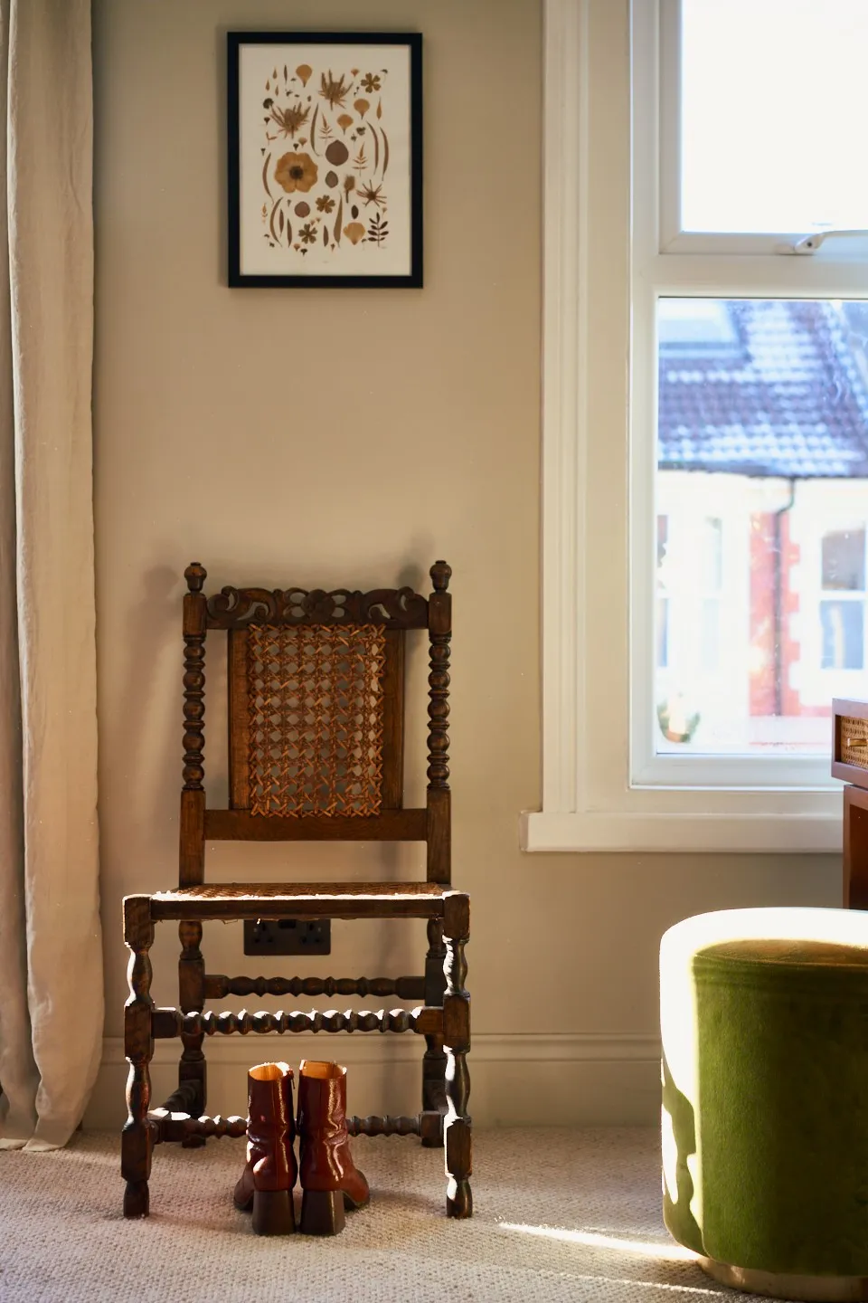 Zoomed in photo in a bedroom, focusing on vintage wooden chair with woven back and seat in front of a beige wall, brown leather boots placed on the floor beneath it, and a botanical print hanging above.