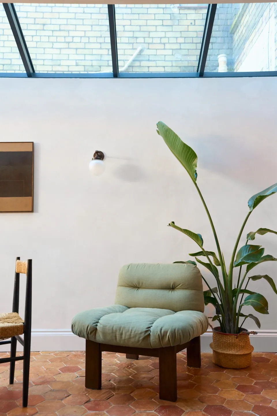 Interior space of an extension, focusing on the materiality and lighting. In the scene there's an accent armchair, an indoor plant in the background and a dining chair on the left. Above, there's a big glass roof. 