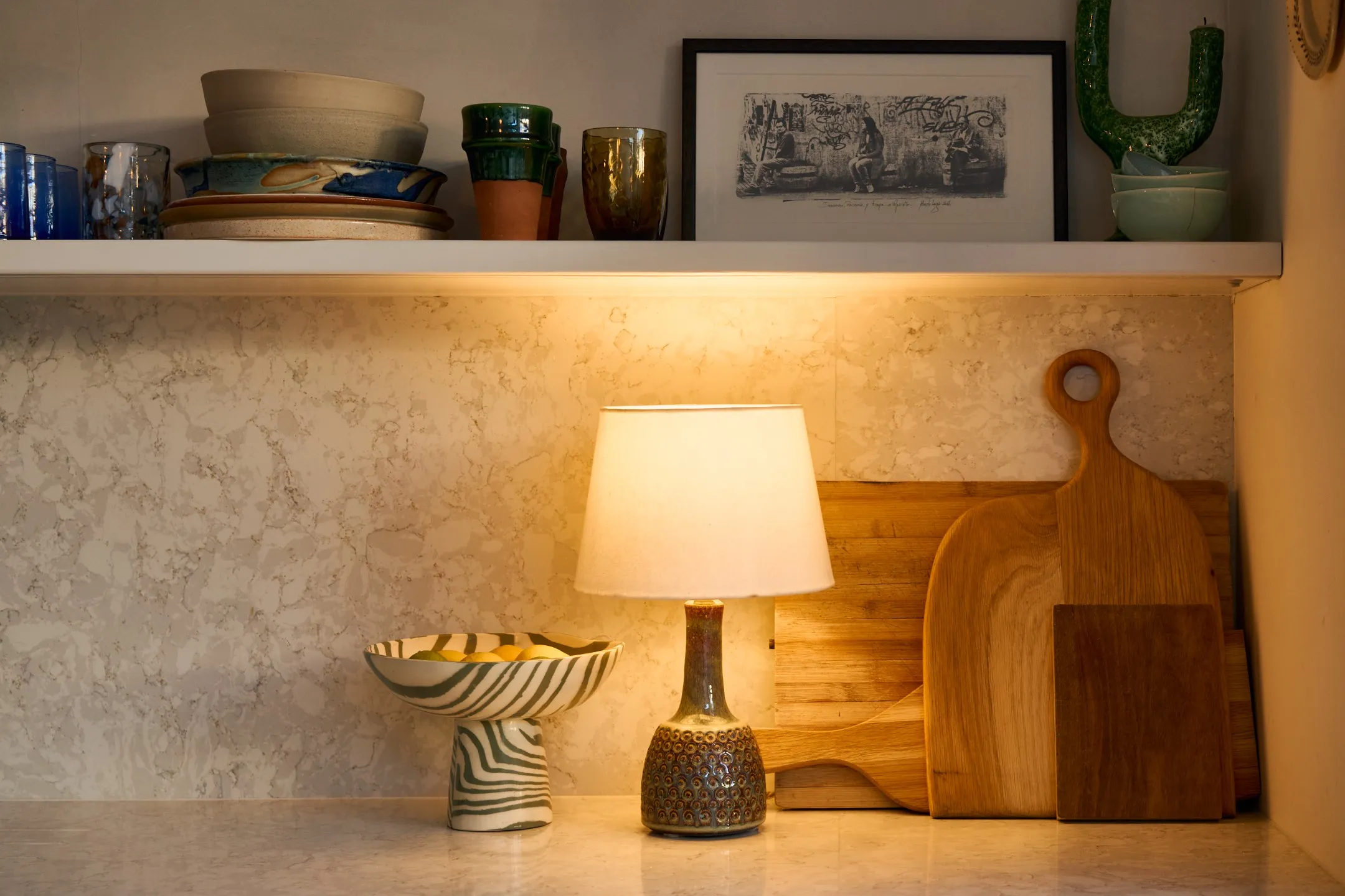 Table lamp with a patterned ceramic base next to a striped pedestal bowl with lemons and wooden cutting boards against a marble backsplash under a white shelf with dishes and framed artwork.