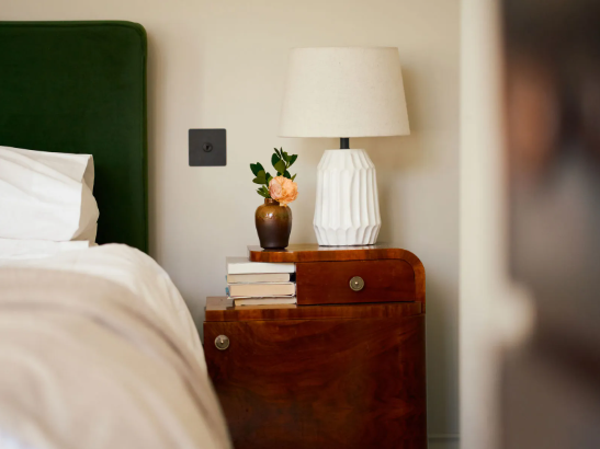 Wooden bedside table with a white lamp, vase with peach flower, and stacked books next to a bed.