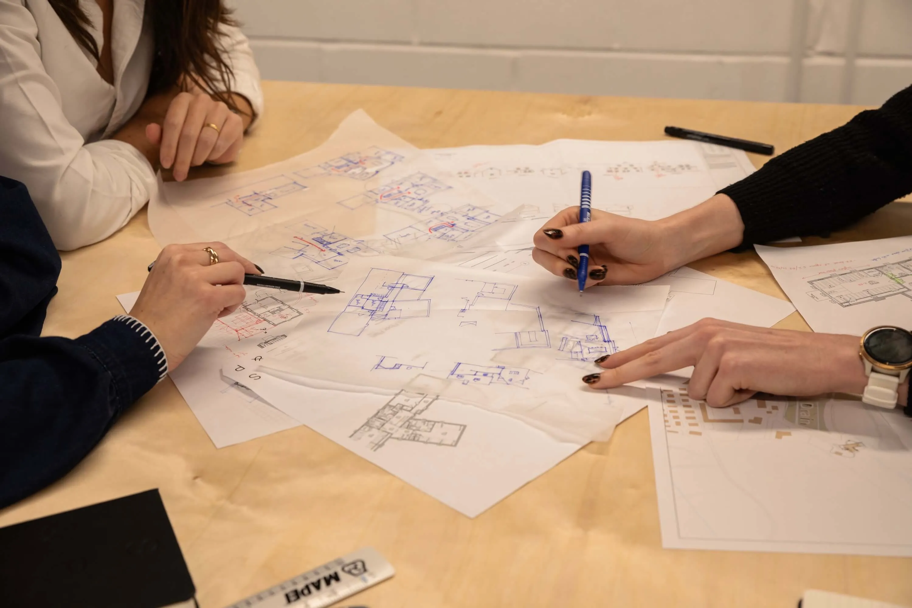 Two people collaborate on architectural floor plans spread on a wooden table, marking details with pens.