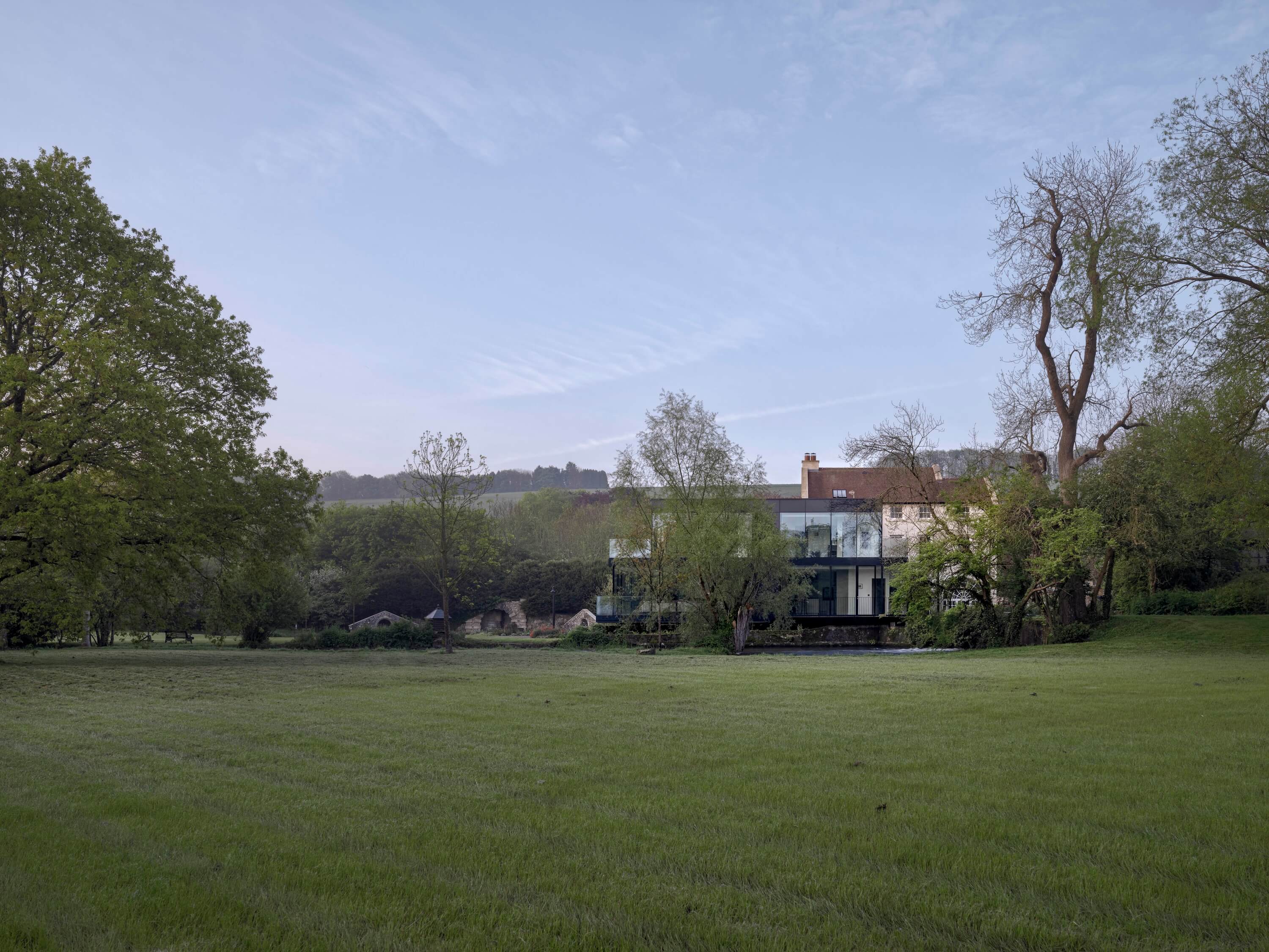 Modern glass and traditional house surrounded by trees with a wide green grassy lawn in front under a blue sky.
