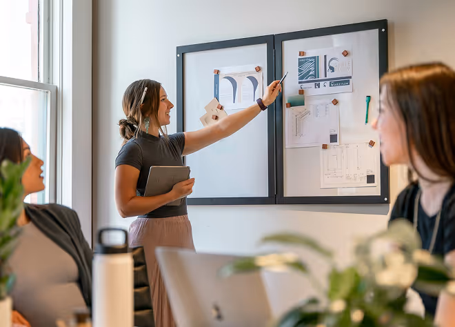 Woman pointing to pinned documents on a wall while holding a tablet, with two women seated and listening in a meeting room.