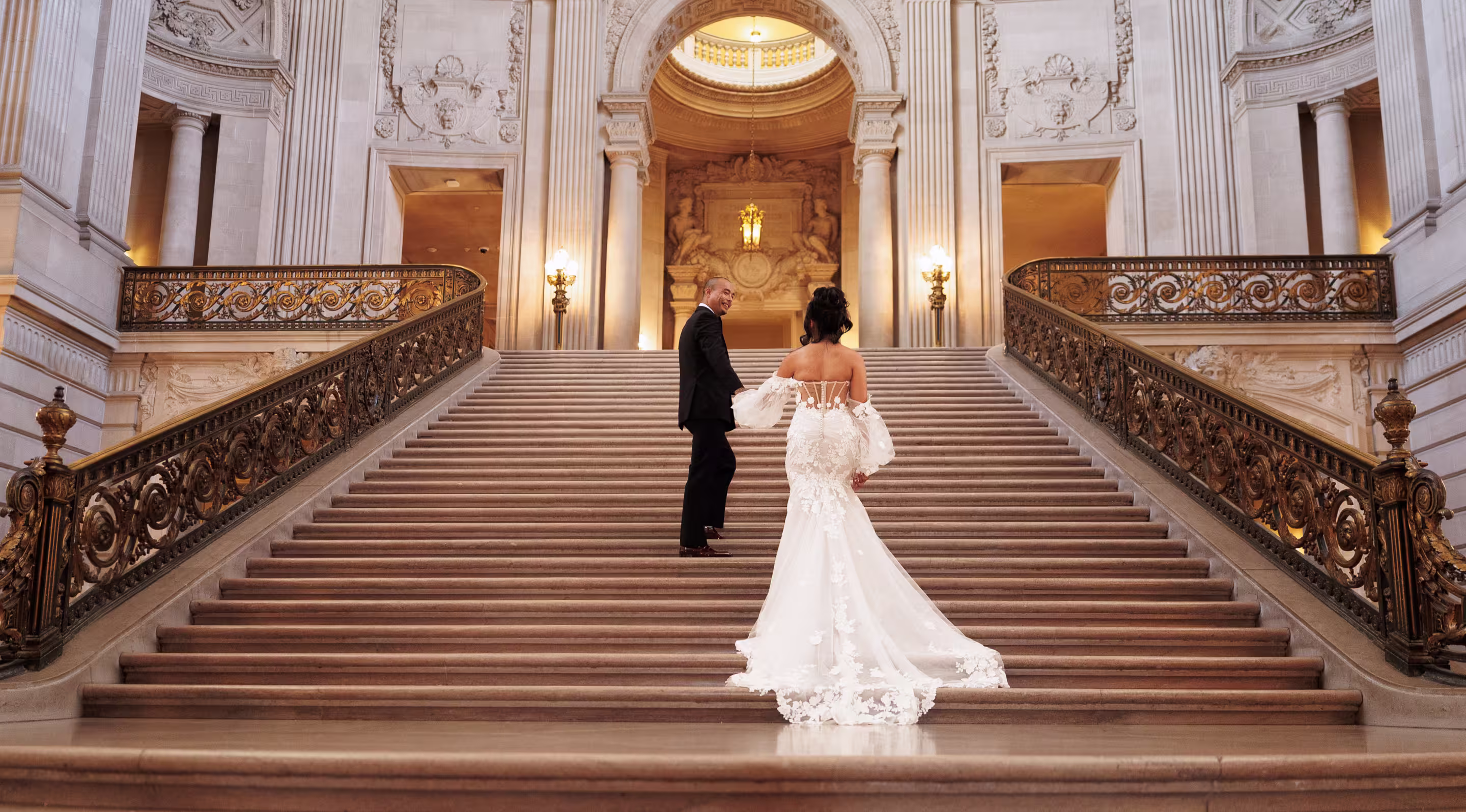 Bride and groom walking up the grand staircase inside San Francisco City Hall during an elegant wedding portrait session, bride in lace gown holding groom's hand, warm architectural lighting and historic marble interior