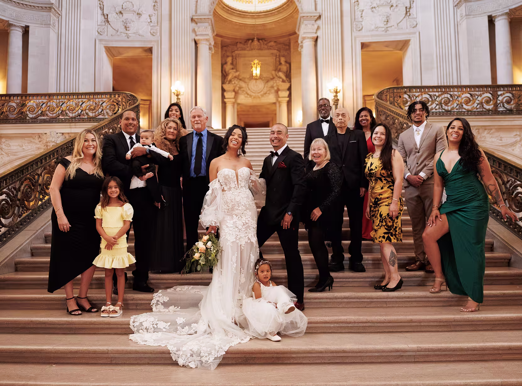 Wedding couple posing with family on the grand staircase at San Francisco City Hall, joyful group portrait with elegant historic architecture and warm light