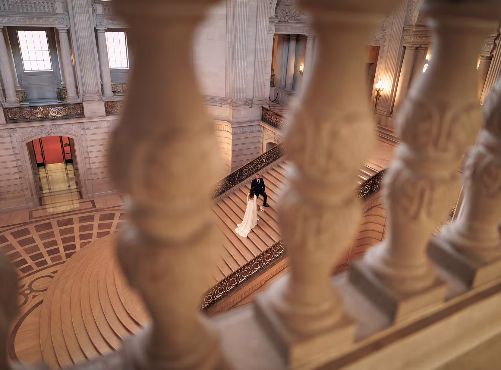 Bride and groom walking on the grand staircase at San Francisco City Hall, photographed through marble balustrades for an elegant architectural perspective