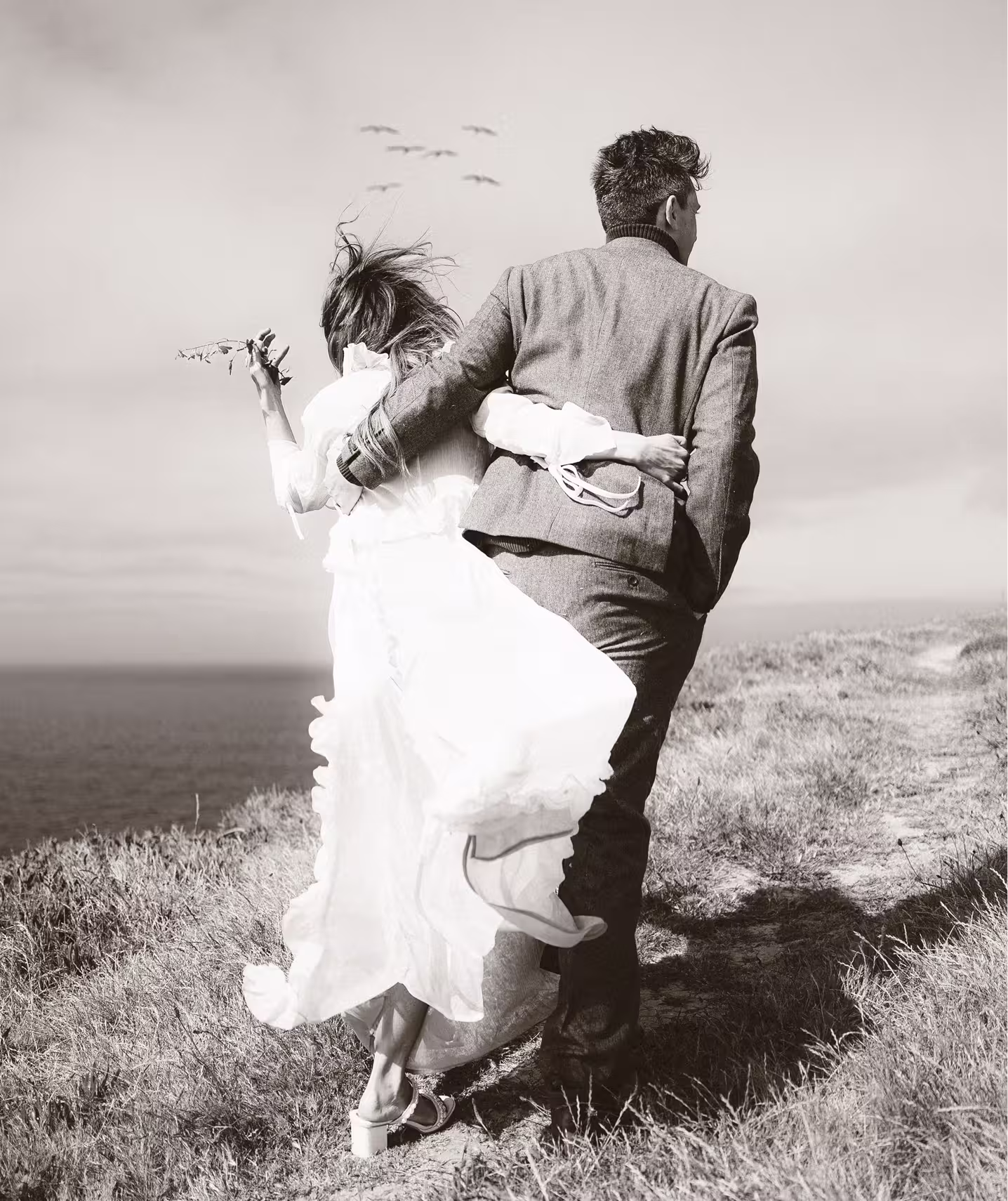 Black and white photo of a wedding couple embracing on a windswept coastal cliff, bride’s dress blowing in the ocean breeze