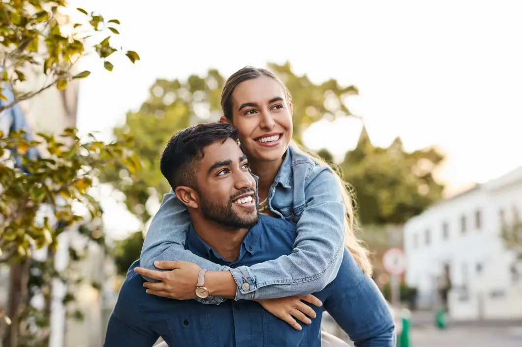 Smiling young woman hugging a man from behind outdoors with trees and buildings in the background.