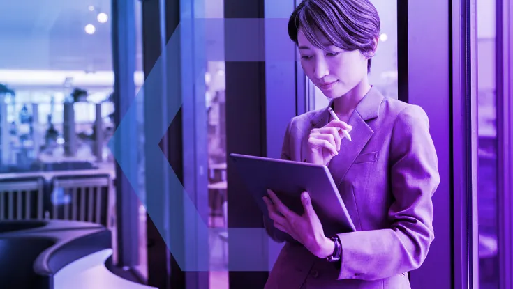 A professional businesswoman in a suit stands in a modern office, focused on her work on a tablet with a stylus