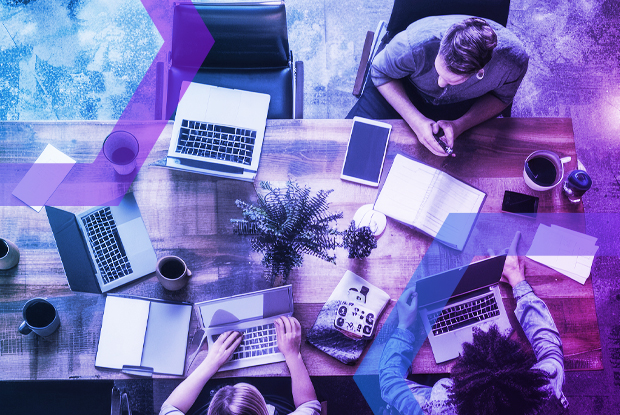 Top-down shot of a diverse team working together in a creative office space with multiple laptops and a tablet. The scene is filtered with a vibrant blue and purple gradient, evoking a sense of modern technology and cybersecurity