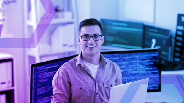 A smiling IT professional holding a laptop in an office with multiple monitors displaying code. The image symbolizes the privacy and control security teams maintain with Menlo Browsing Forensics, as all forensic data is sent directly to their own storage systems.