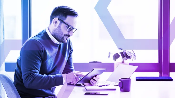 A man with glasses wearing a blue sweater sits at a desk, looking at a tablet he is holding. In front of him are a laptop and two large computer monitors displaying code.