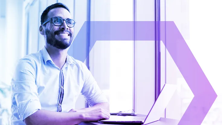 smiling man with glasses sits at his laptop in a bright, modern office. His confident and relaxed expression represents the ease and efficiency of the 'Simplified, Agentless Zero Trust Deployment' described in the text