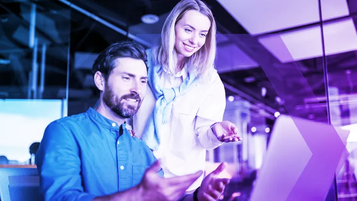 A man sits at a desk while his female colleague stands beside him, leaning over to point something out on a laptop screen. Both are focused and engaged, working together in an office environment with a blue and purple technological color overlay.